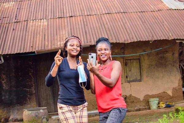 Two Happy African women, ladies or friends happily look into a smart phone outside a village mud house