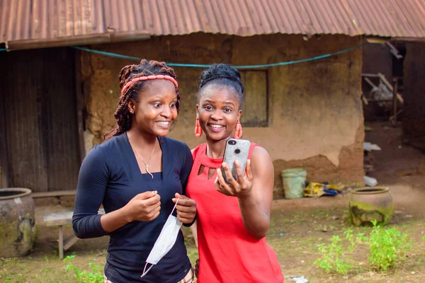 Two Happy African women, ladies or friends happily look into a smart phone outside a village mud house