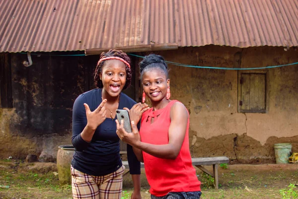 Two Happy African women, ladies or friends happily look into a smart phone outside a village mud house