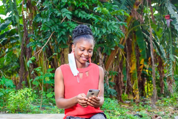 A happy woman or lady with nose mask, wearing a red dress, sitting and using a smart phone in a farm with banana trees behind her