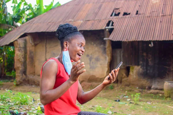 A happy woman or lady with nose mask, wearing a red dress, sitting and using a smart phone outside a village mud house
