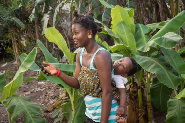 Sırtında bir bebekle güzel bir Afrikalı anne ve muz çiftliğinde dururken elinde akıllı bir telefon. 