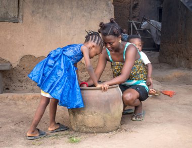  Çocuklarıyla birlikte köyün çamurlu evinin önünde çömelmiş büyük bir çömleğin ya da kabuğun yanında eğilmiş Afrikalı bir anne. 
