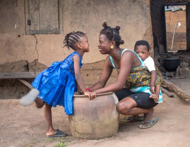  Çocuklarıyla birlikte köyün çamurlu evinin önünde çömelmiş büyük bir çömleğin ya da kabuğun yanında eğilmiş Afrikalı bir anne. 