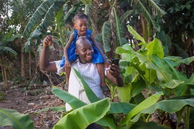 Mutlu Afrikalı baba elinde telefonla muz çiftliğinde kızını taşıyor.
