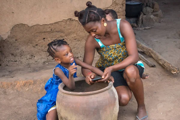 An African mother with her children bending low beside a big clay pot or calabash in front of a village mud house  An African mother with her children bending low beside a big clay plot in front of a village mud house with a mobile phone