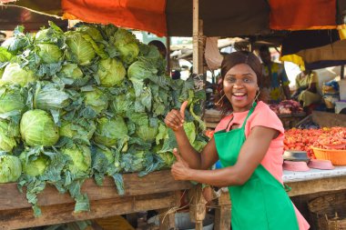 Happy African business woman or female trader wearing a green apron and doing thumbs up gestures while standing at her vegetable stall in a market place