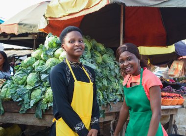 Two happy African business women or female traders wearing colorful aprons and standing together in front of their vegetable stall in a market place