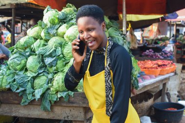 Happy African business woman or female trader wearing a yellow apron and making a call with a smart phone while standing at her vegetable stall in a market place