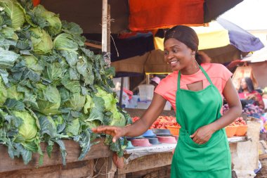 Happy African business woman or female trader wearing a green apron and tending to her goods of vegetables in a market place