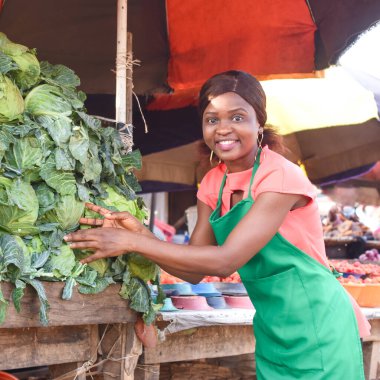 Happy African business woman or female trader wearing a green apron and tending to her goods of vegetables in a market place