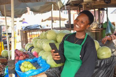 Yeşil önlüklü mutlu bir Afrikalı kadın ya da kadın tüccar ve akıllı bir telefonla yerel bir markette tezgahında duruyor.