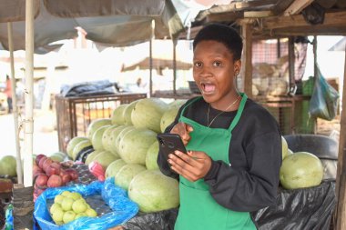 Afallamış bir Afrikalı kadın ya da kadın tüccar yeşil önlüklü ve akıllı telefonlu, yerel bir markette tezgahında duruyor.