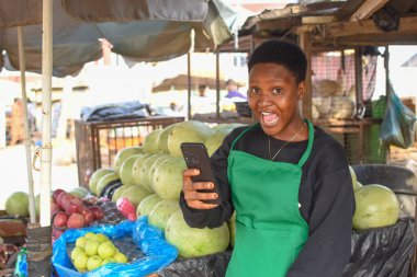Afallamış bir Afrikalı kadın ya da kadın tüccar yeşil önlüklü ve akıllı telefonlu, yerel bir markette tezgahında duruyor.