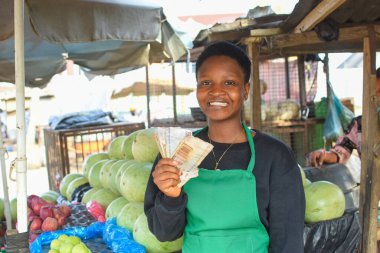 A happy African woman or female trader in green apron, holding some Naira notes or cash while standing at her stall in a local market