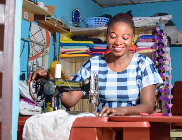 A happy female African Nigerian tailor, fashion designer, business woman or entrepreneur sitting down with while working with a sewing machine in a tailoring shop