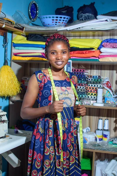 A happy female African Nigerian tailor, fashion designer, business woman or entrepreneur with a tape measure around her neck and looking at the camera in a tailoring shop