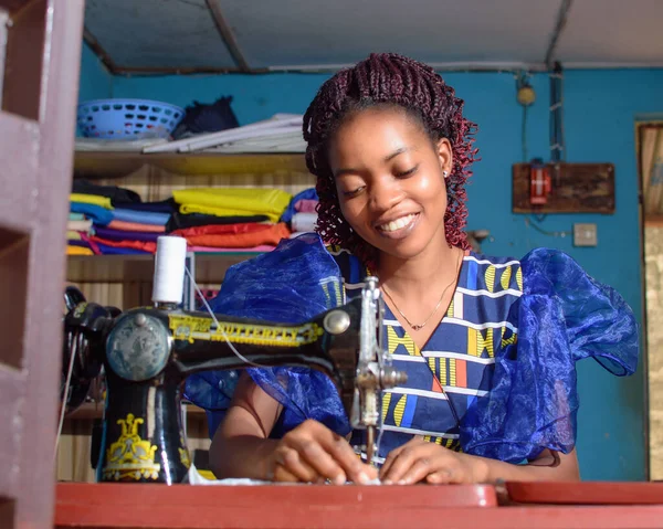 A happy female African Nigerian tailor, fashion designer, business woman or entrepreneur sitting down with while working with a sewing machine in a tailoring shop