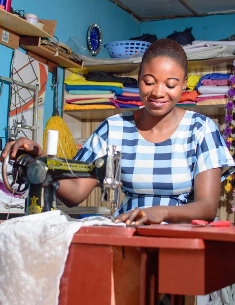 A happy female African Nigerian tailor, fashion designer, business woman or entrepreneur sitting down with while working with a sewing machine in a tailoring shop