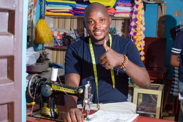 African Nigerian male tailor, fashion designer or business man working with a sewing machine in a tailoring shop and doing thumb up gesture