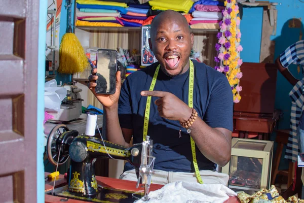 A happy African Nigerian male tailor, fashion designer or business man pointing to a smart phone in his hand while working with a sewing machine in a tailoring shop