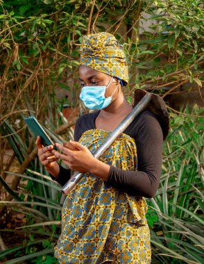 A beautiful African female farmer with nose mask, checking her smartphone with a hoe hanging from her shoulder in her pineapple farm