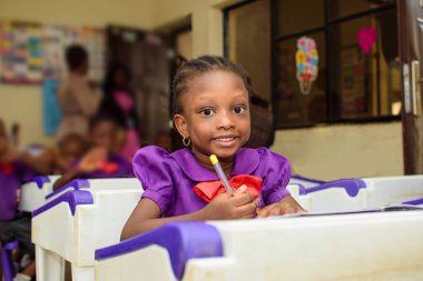 African girl child wearing a purple school uniform, studying and writing with a pen in a book while sitting down in a classroom to study for excellence in her education
