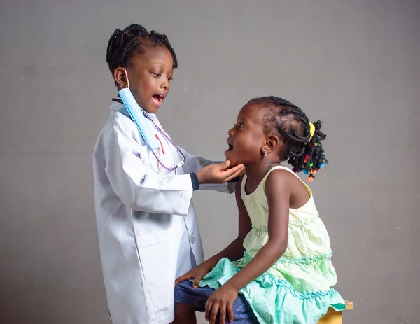 African Nigerian girl child with nose mask and stethoscope, playing medical expert like doctor, nurse and dentist with kid patient sitting in front of her