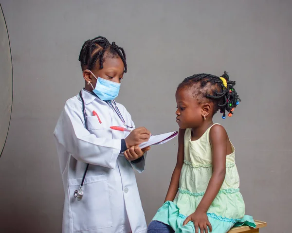 African Nigerian girl child with nose mask and stethoscope, playing medical expert like doctor and nurse with kid patient sitting in front of her