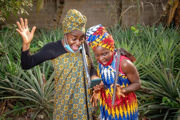 Two beautiful African female farmers with nose mask, looking happily into a smartphone with a hoes hanging from their shoulders in a pineapple farm