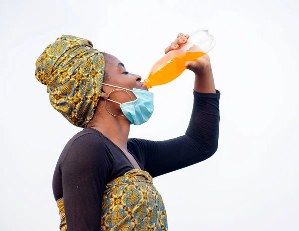 A beautiful African lady drinking a yellow liquid content from a transparent bottle with her nose mask below her jaw