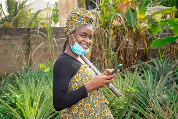 A beautiful African female farmer with nose mask, checking her smartphone with a hoe hanging from her shoulder in her pineapple farm