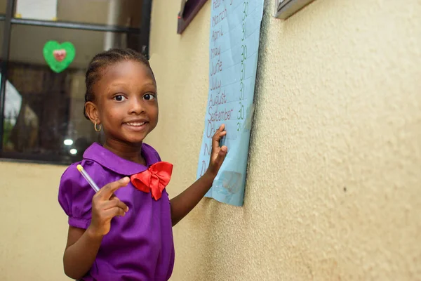 African girl child wearing a purple school uniform and standing in a classroom with a pen in her hand to study for excellence in her education