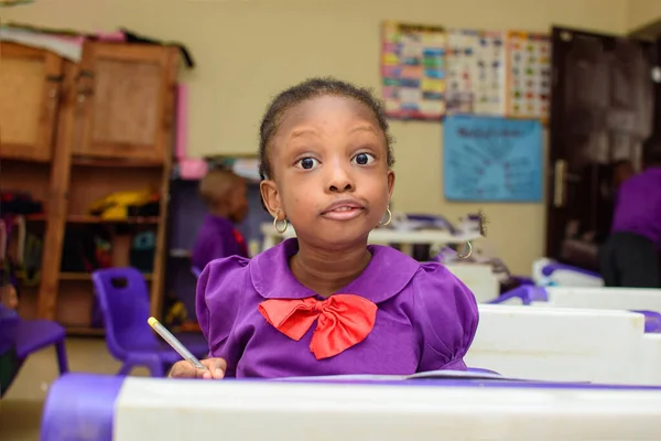 African girl child wearing a purple school uniform, studying while writing with a pen in a book and sitting down in a classroom to study for excellence in her education