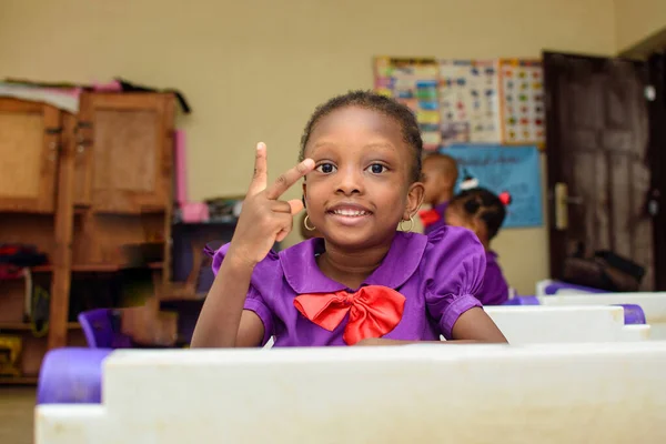 African girl child making happy gestures with fingers, wearing a purple school uniform while sitting at a desk and studying in a classroom for excellence in her education