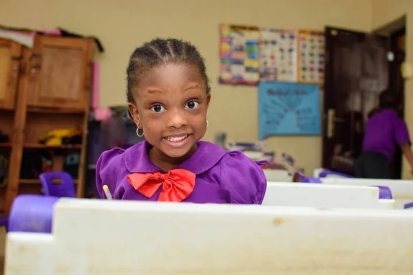 African girl child wearing a purple school uniform, studying and writing with a pen in a book while sitting down in a classroom to study for excellence in her education