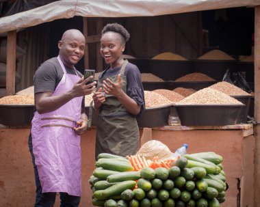 A male and female African Nigerian traders, sellers, business persons or shop owners, having aprons on their body and happily looking at the camera while holding a smart phone in a market