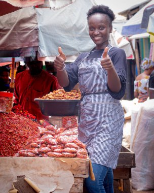 An African Nigerian female trader, seller, business woman or entrepreneur with an apron, happily doing thumbs up gestures to attract customers to her stall in a market