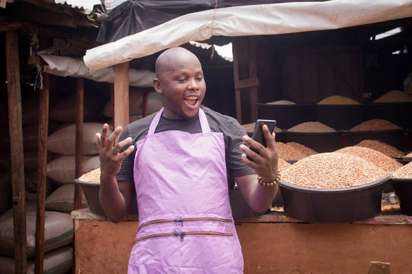 An African Nigerian male trader, seller, business man or shop owner, having an apron on his body and happily looking into the smart phone he is holding in front of his goods in a market