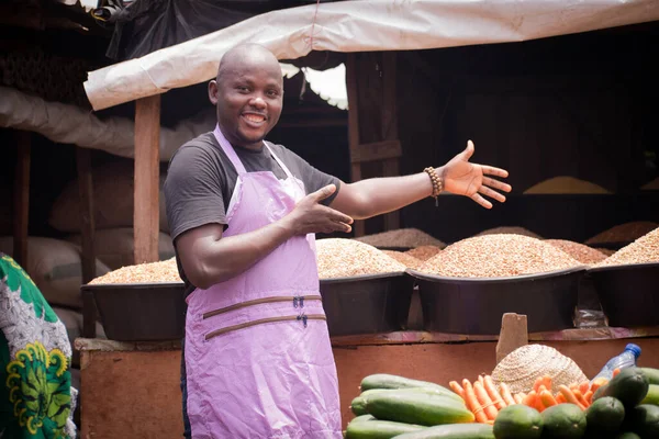 An African Nigerian male trader, seller, business man or shop owner, having an apron on his body and attracting potential customers to his displayed goods in a market