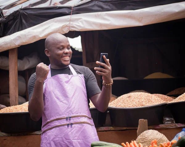 An African Nigerian male trader, seller, business man or shop owner, having an apron on his body and amazed as he stares into the smart phone in his hand in a market