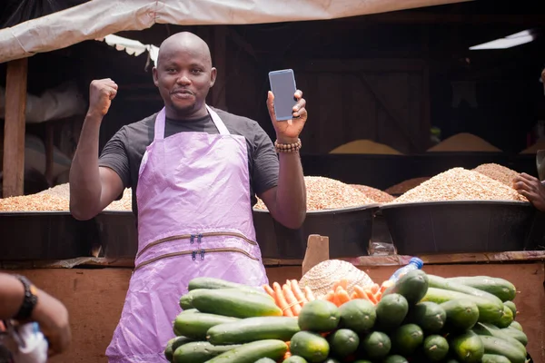 An African Nigerian male trader, seller, business man or shop owner, having an apron on his body, happily jubilating and displaying a smart phone he his holding in a market