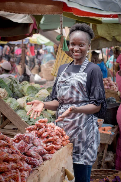 An African Nigerian female trader, seller, business woman or entrepreneur with an apron, happily showing her displayed goods to attract potential customers in a market