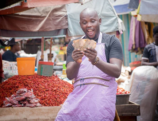An African Nigerian male trader, seller, business man or entrepreneur with an apron, happily showing and counting the money from his sales in a market