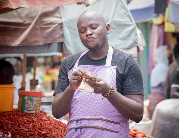 An African Nigerian male trader, seller, business man or entrepreneur with an apron, happily showing and counting the money from his sales in a market