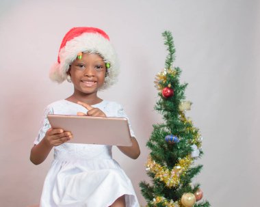 Portrait of a cute female African girl child with a smart phone or tablet, wearing a Santas cap with a Christmas tree beside her 