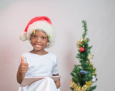 Portrait of a cute female African girl child with a smart phone or tablet, wearing a Santas cap with a Christmas tree beside her 