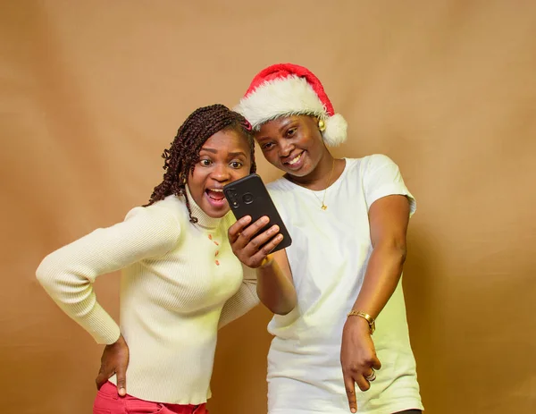 Two African ladies, sisters or friends happily looking at a smart phone while one of them have a Christmas cap on her head