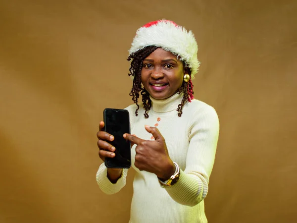 An African lady pointing to the smart phone in her hand and also have a Christmas cap on her head
