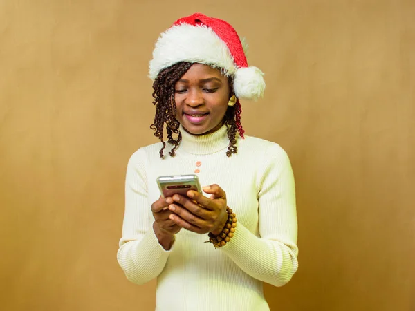 An African lady looking into the smart phone in her hands and also have a Christmas cap on her head